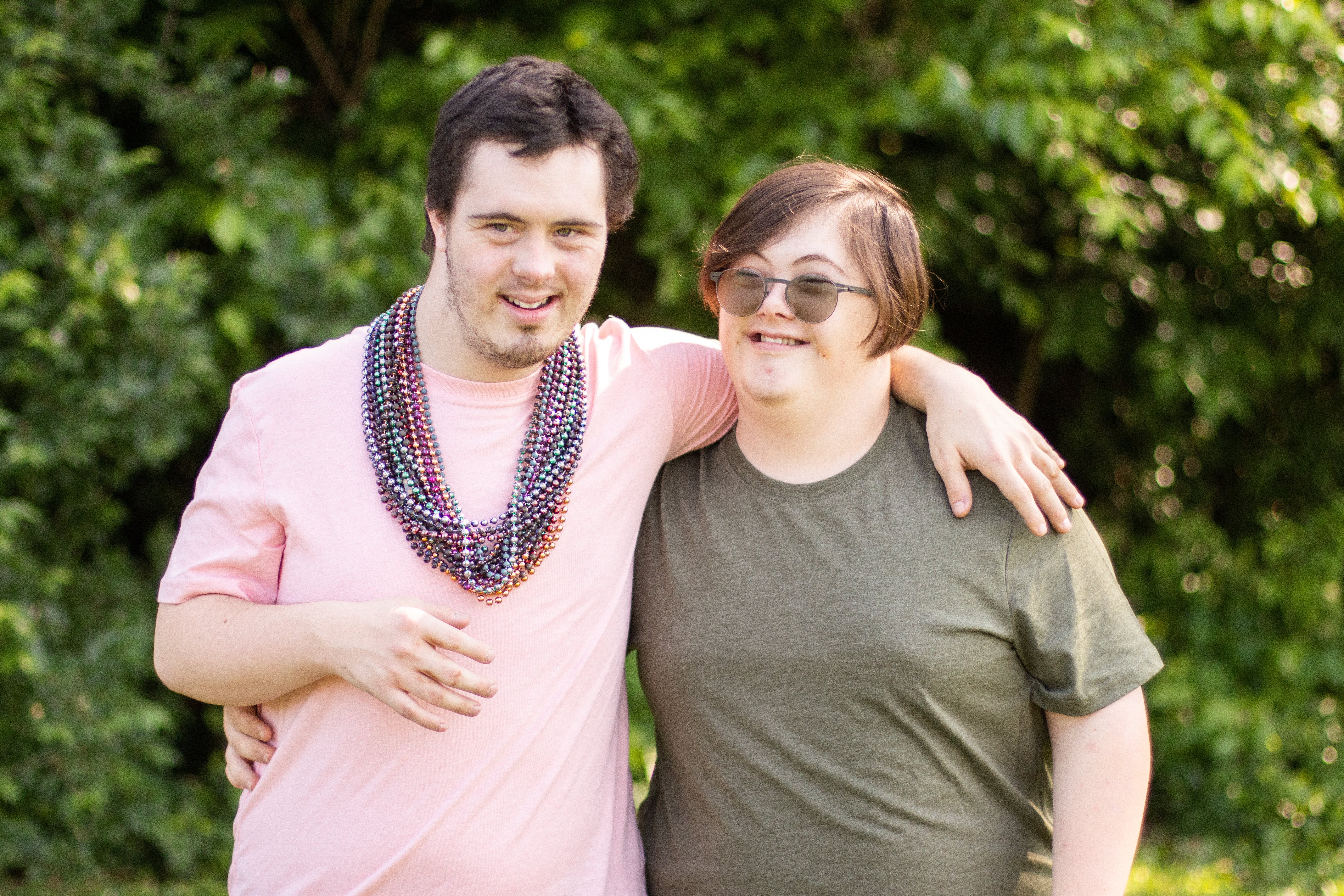 Two Young Adults Smile and Stand Next to Each Other with Arms over Shoulders with Trees in Background